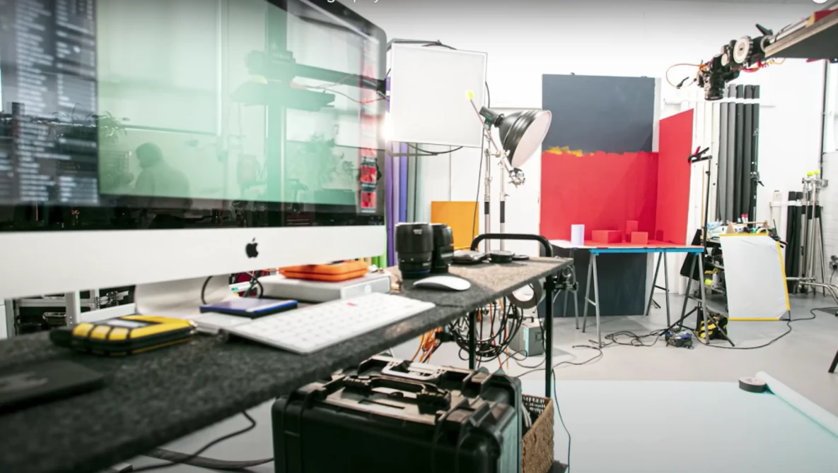 Professional photography studio workspace with Mac monitor, lighting equipment, and red backdrop panels.