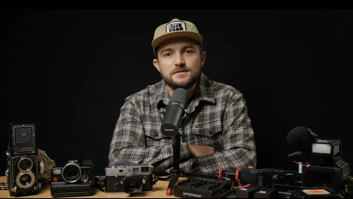 Photographer at desk with vintage and modern cameras speaking into microphone against dark background.