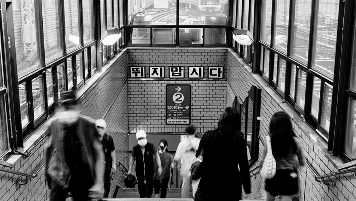 Blurred commuters moving through a subway station stairwell with Korean signage and brick walls.