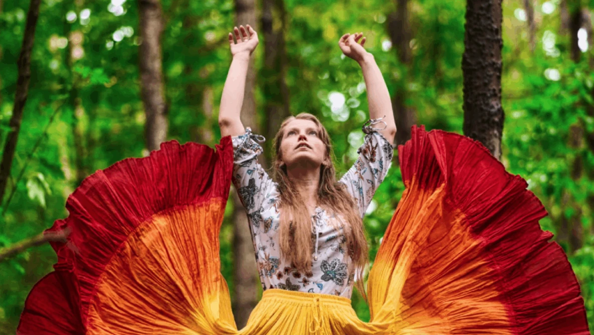 Woman with arms raised wearing flowing red and yellow fabric against green forest backdrop.