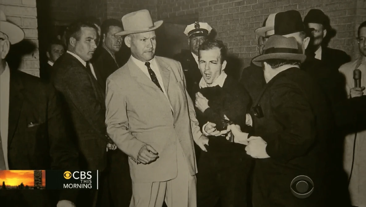 Men in suits and fedoras gathered in a brick corridor, one in a light tan coat being escorted by officials.