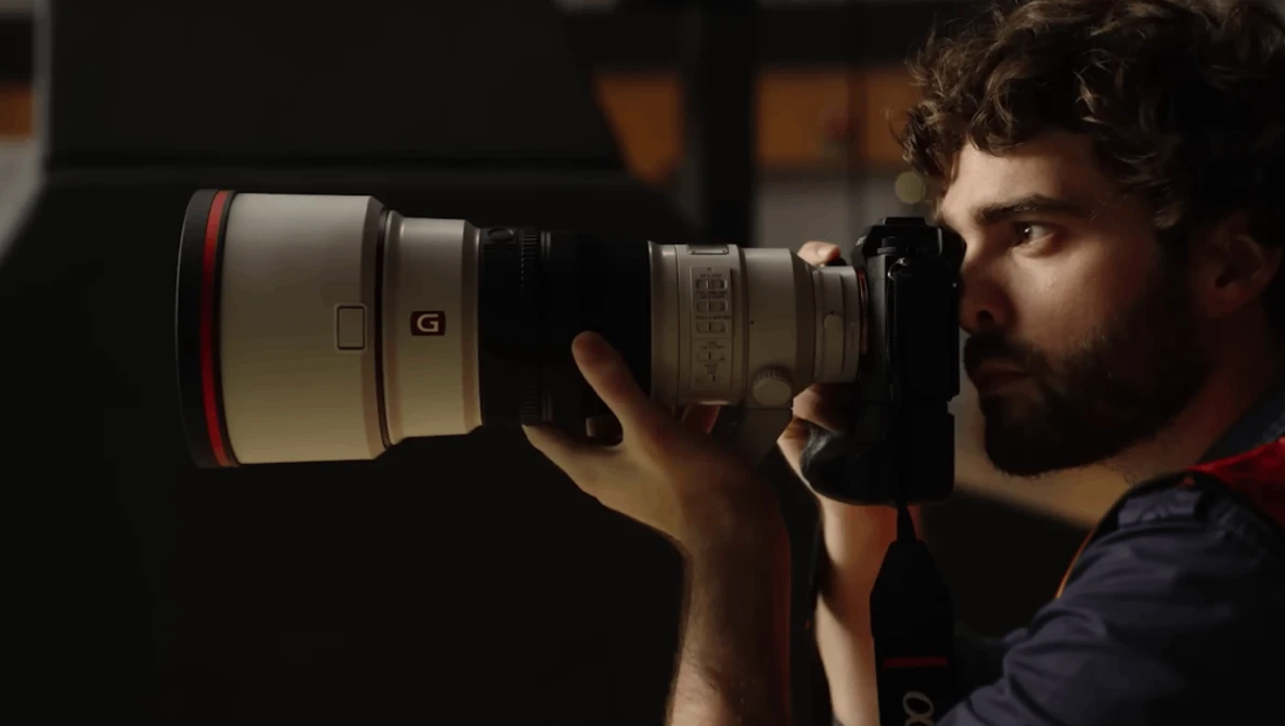 Photographer holding a telephoto lens camera up to his eye, composing a shot in dramatic side lighting.