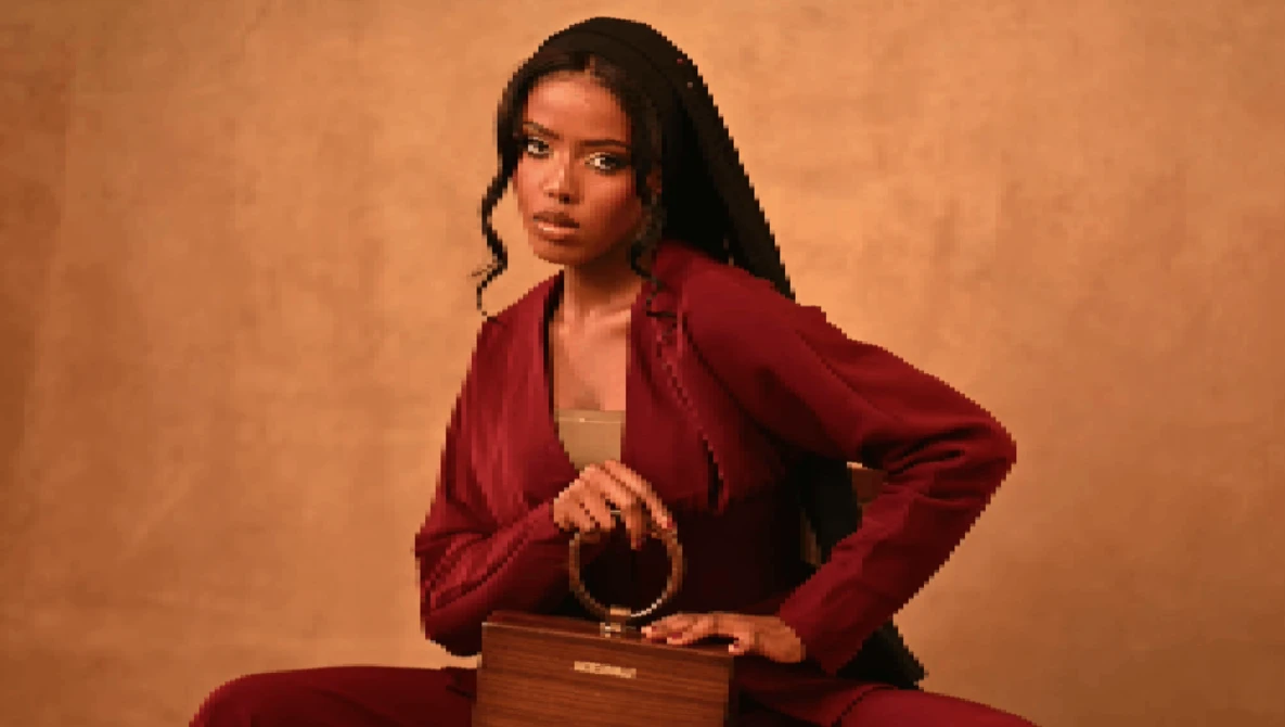 Woman in burgundy blazer posing with wooden box against warm tan backdrop.