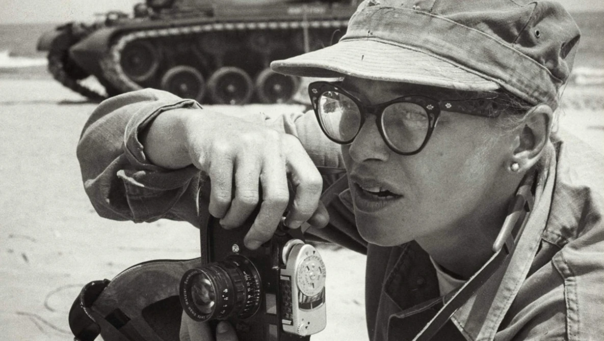 Photographer wearing glasses and cap looking through rangefinder camera with military tank blurred in background.