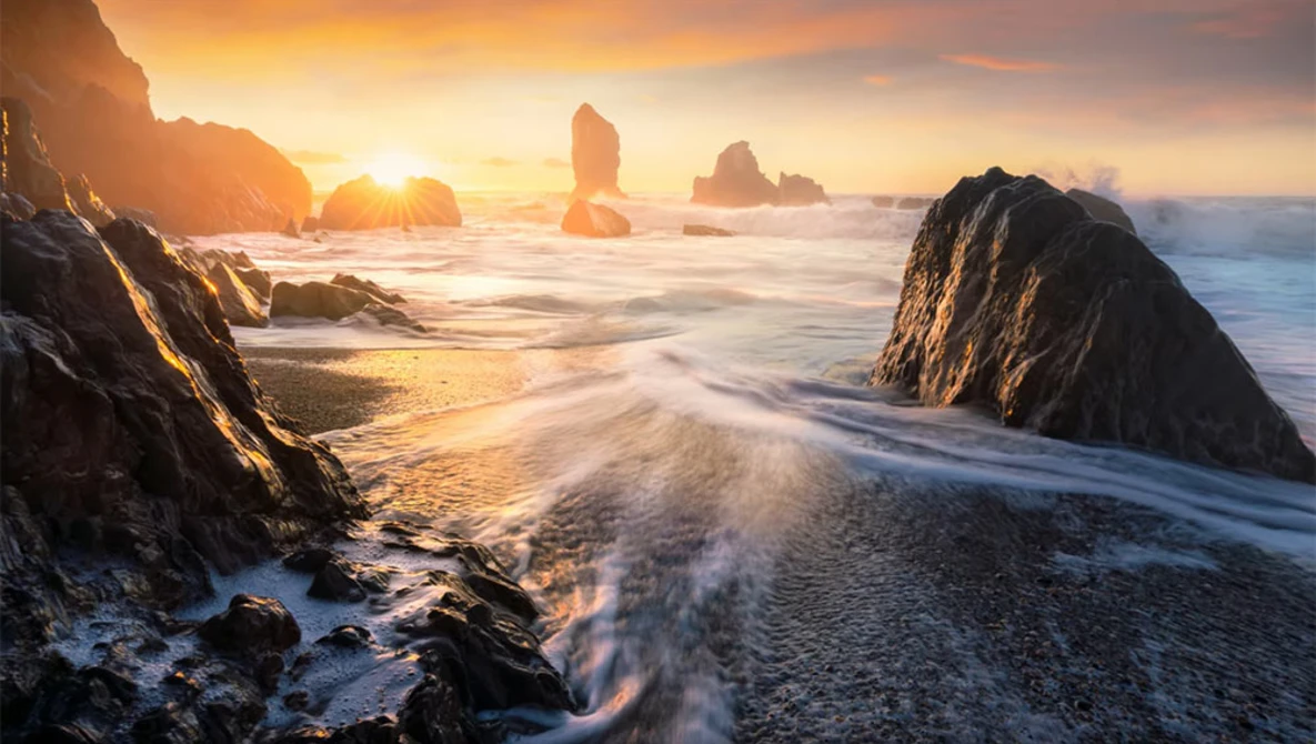Rocky sea stacks silhouetted against golden sunrise over sandy beach with breaking waves.