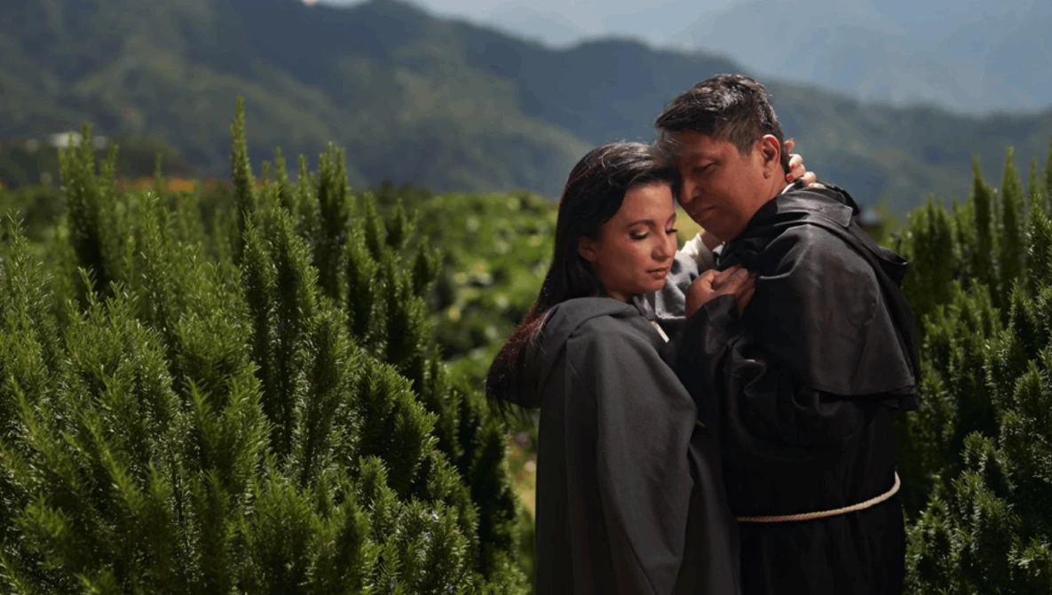 Two people in dark clothing embrace among tall green cypress trees with mountains visible in the soft-focused background.