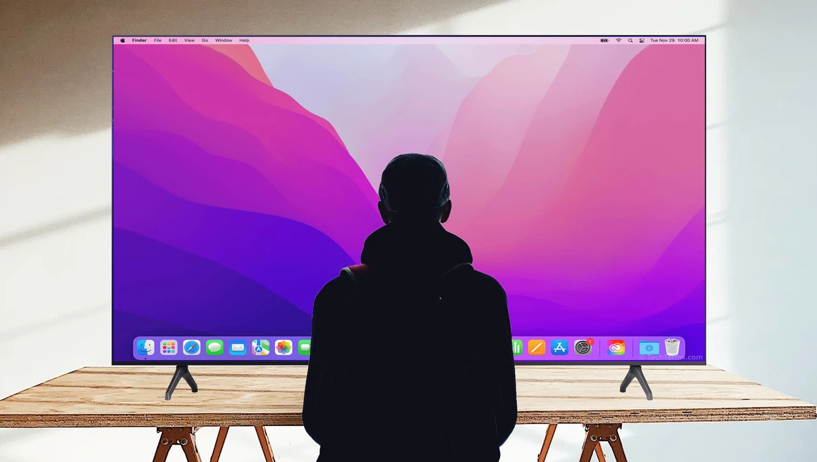 Person sitting at wooden desk viewing a large monitor displaying a vibrant pink and purple gradient wallpaper.