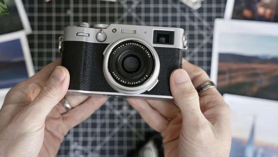 Hands holding a silver and black rangefinder camera with fixed lens against a plaid background.