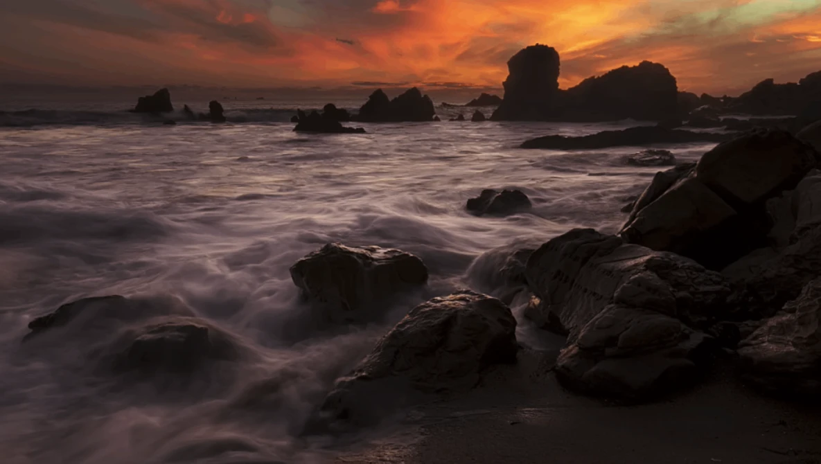 Rocky coastline with crashing waves silhouetted against a dramatic orange and gold sunset sky.