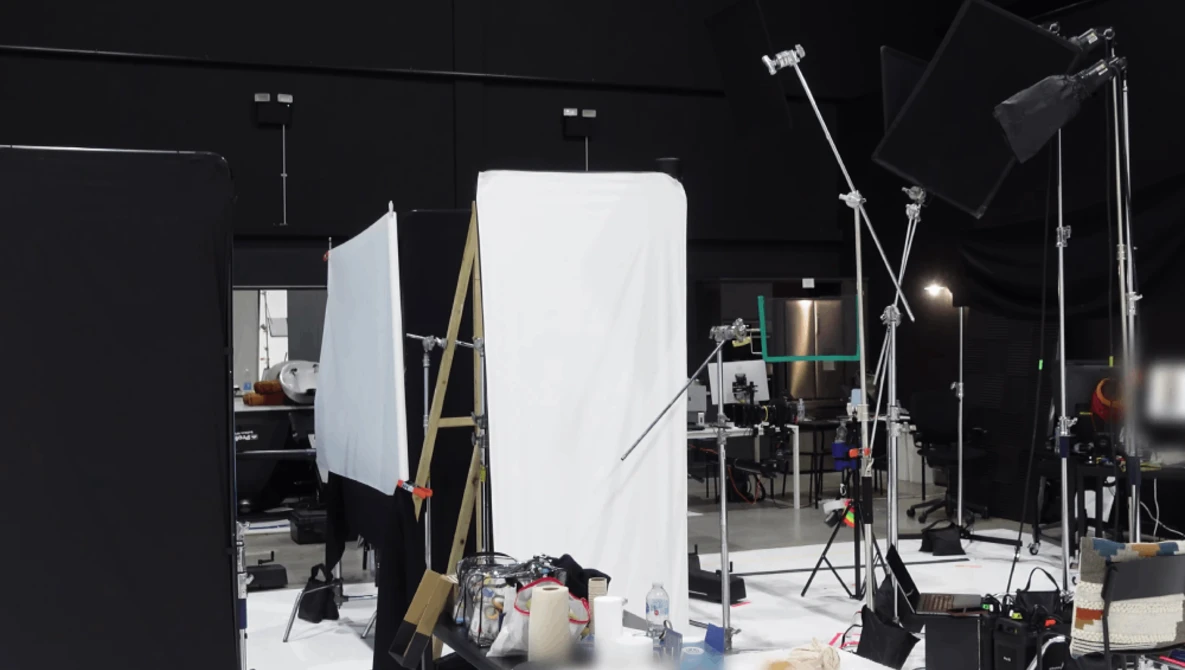 Professional photography studio setup with large white reflector panel, multiple light stands, and softbox lighting on black walls.