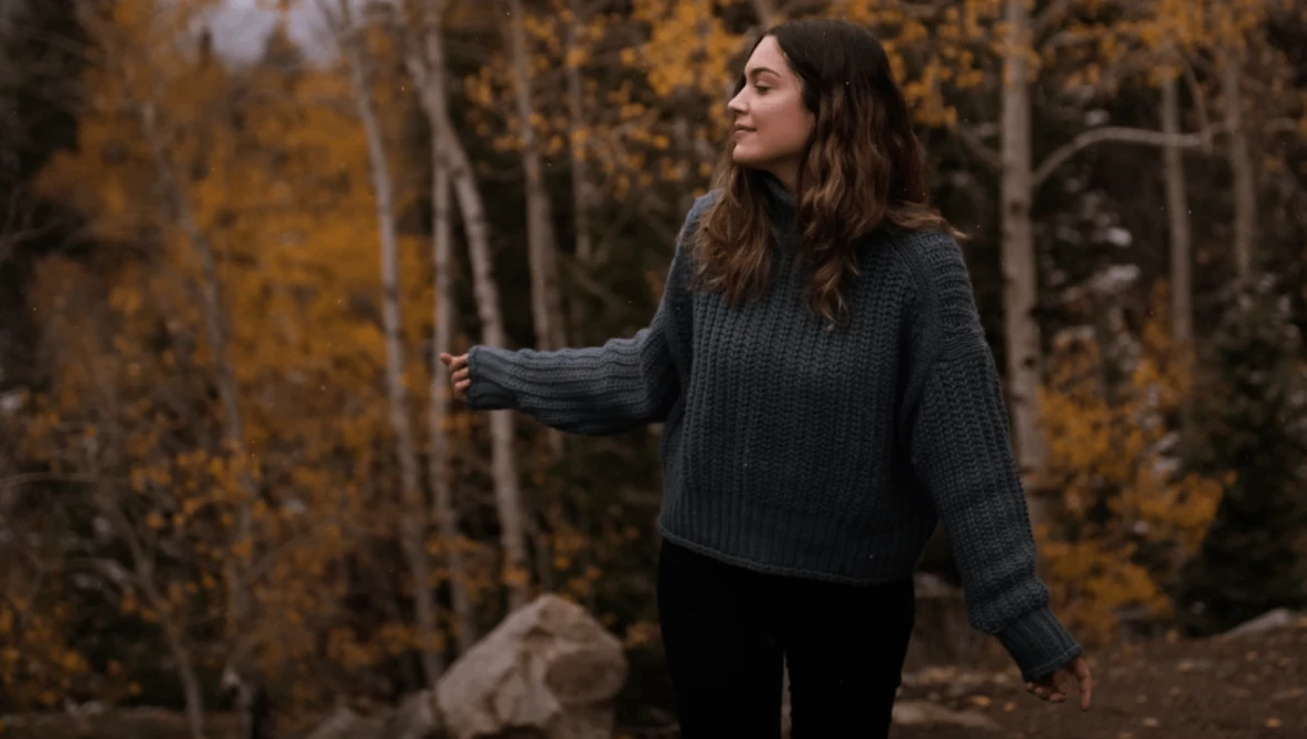 Woman in dark sweater standing in forest with golden autumn birch trees behind her.
