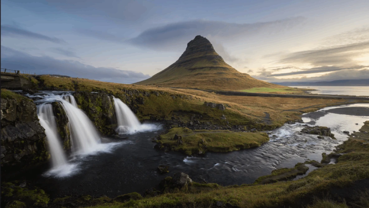 Twin waterfalls cascade into a dark river with a distinctive cone-shaped mountain rising behind moss-covered terrain.