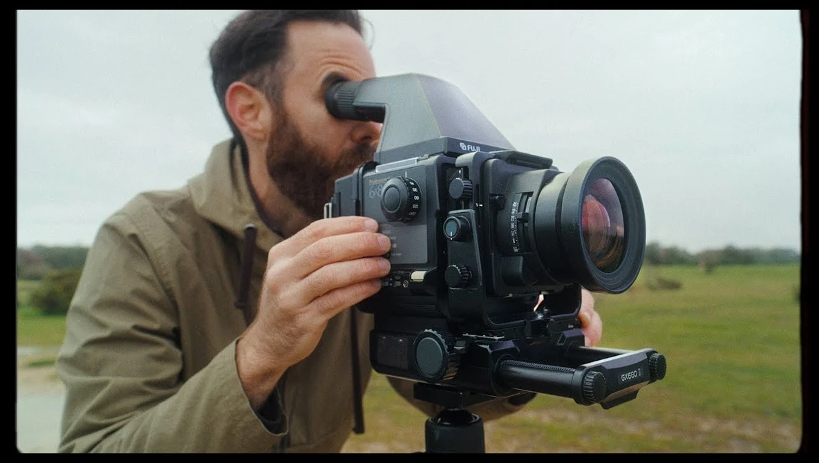 Cinematographer operating a film camera with viewfinder and attached lens in an open field.
