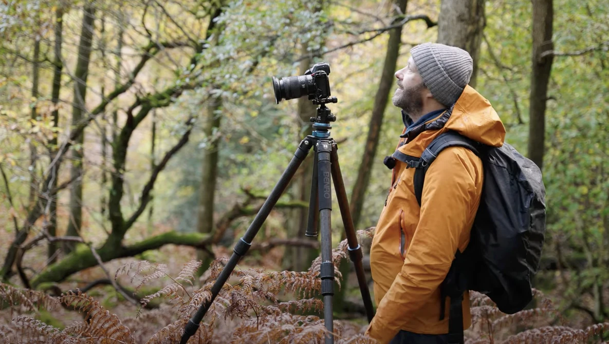 Photographer with camera on tripod in forest setting, wearing orange jacket and gray beanie.