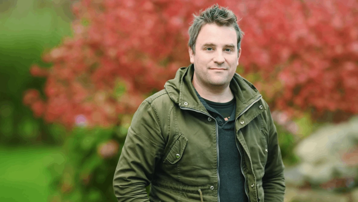 Man in olive jacket standing outdoors with red foliage in soft focus background.