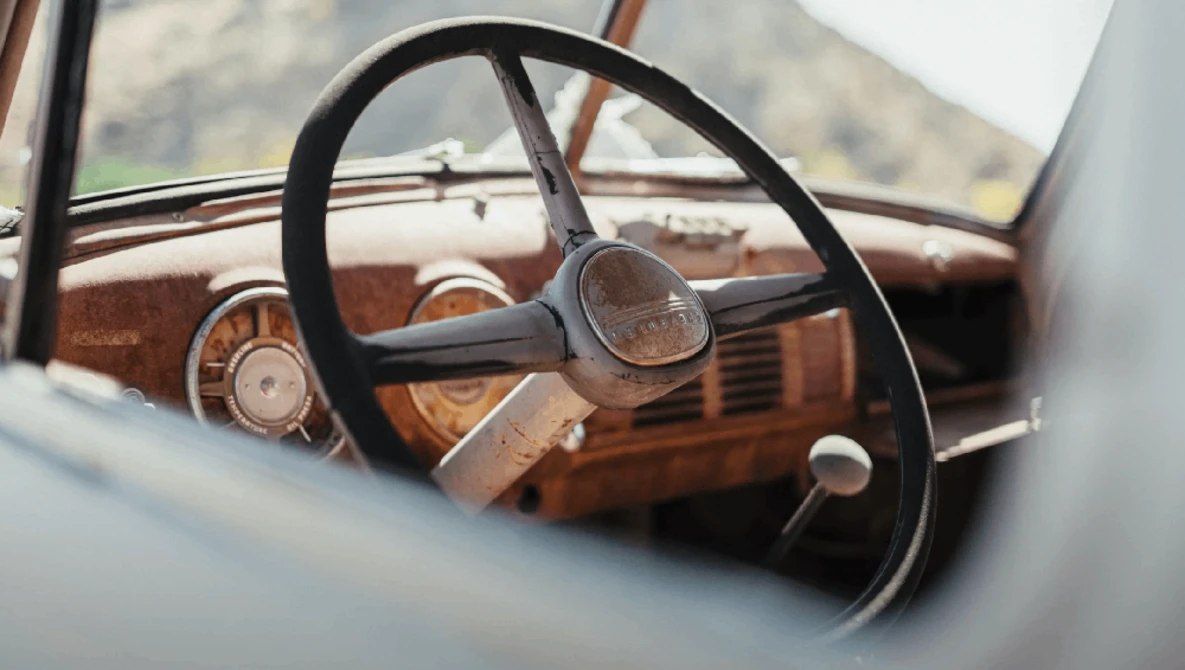 Vintage car interior with worn wooden dashboard and black steering wheel visible through windshield.