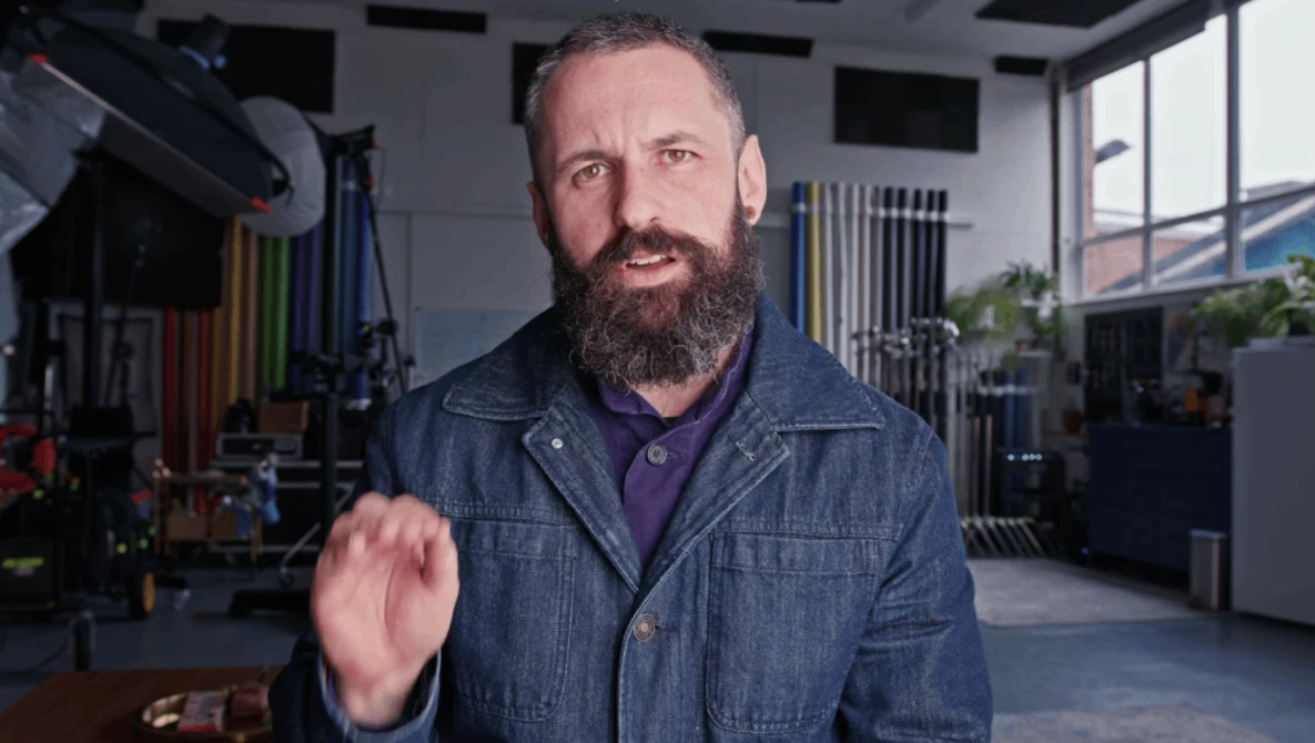 Bearded man in denim shirt gesturing while speaking in a studio workshop.