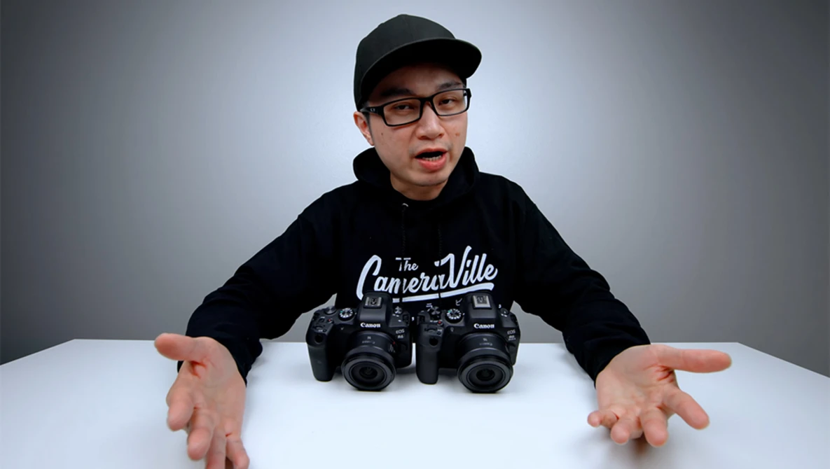 Young photographer wearing glasses and black cap displays two professional camera bodies on white table.