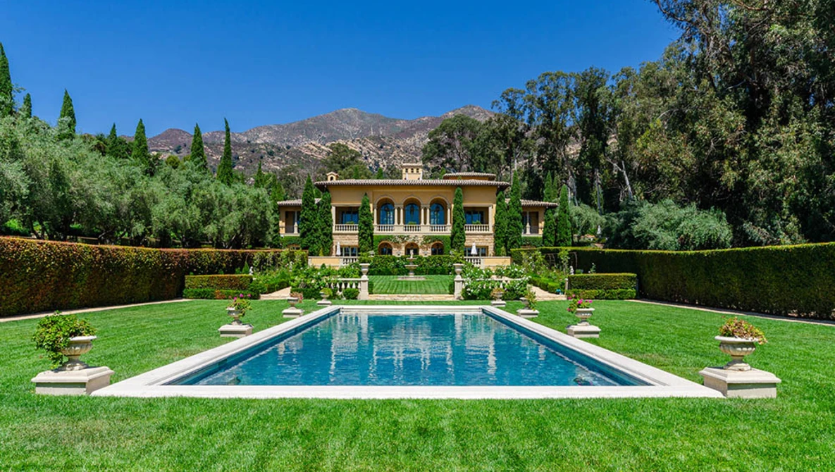 Symmetrical formal garden with rectangular swimming pool, manicured lawn, and yellow villa framed by cypress trees and mountains.