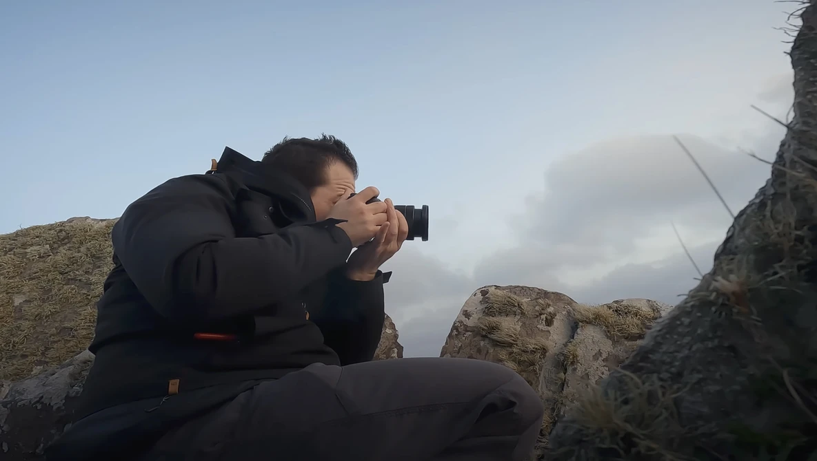 Photographer crouched on rocky terrain, holding a camera to their face while composing a shot against a cloudy sky.