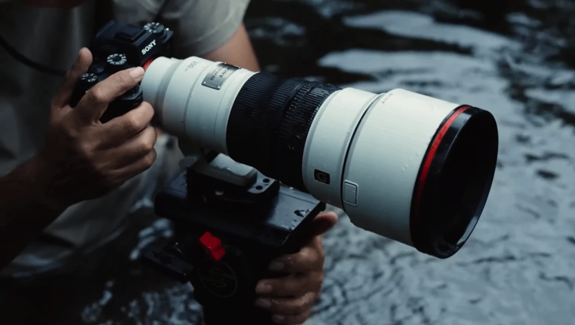 Photographer holding a mirrorless camera with a white telephoto lens featuring a red accent ring.