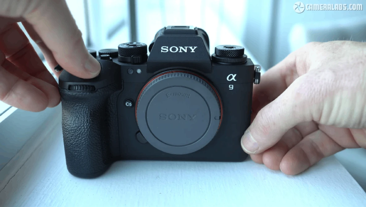 Hands holding a Sony Alpha 9 mirrorless camera body with attached grip against a bright window background.