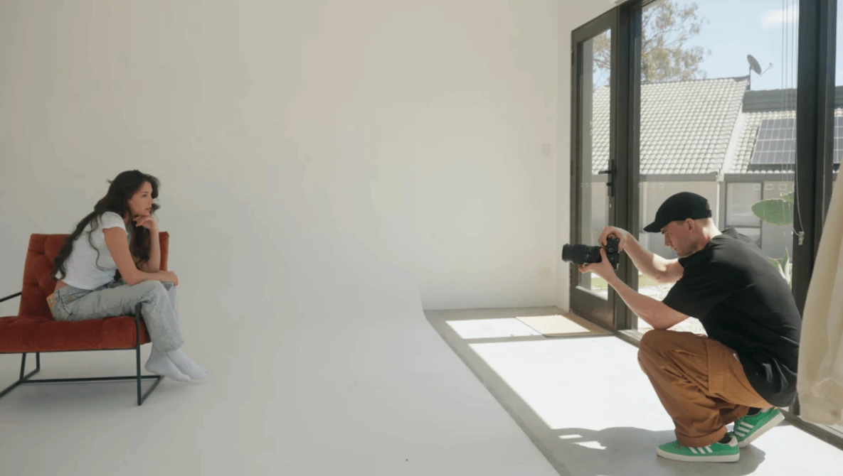 Photographer crouching while shooting a model seated on a red chair in a minimalist white studio.