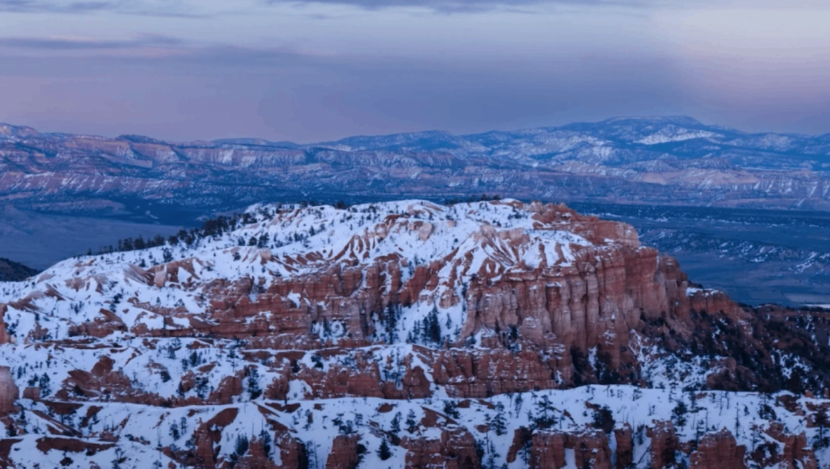 Snow-covered hoodoo formations and ridges in red rock canyon landscape with distant mountains under twilight sky.