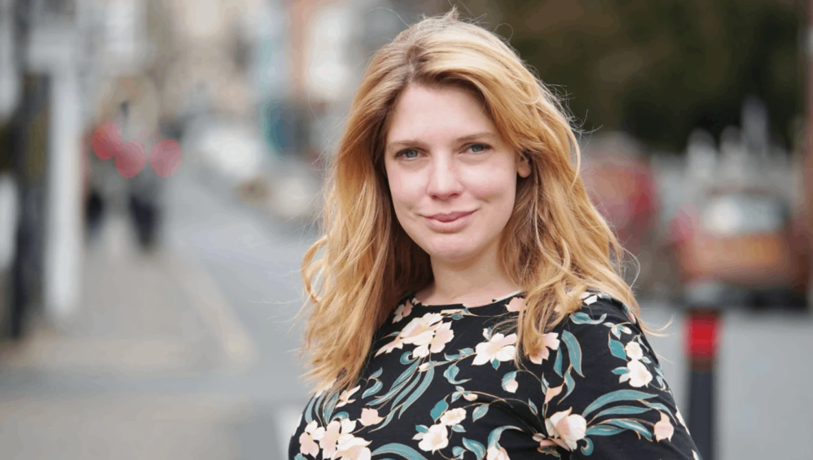 Woman with strawberry blonde hair wearing a floral patterned top, photographed outdoors on a city street.