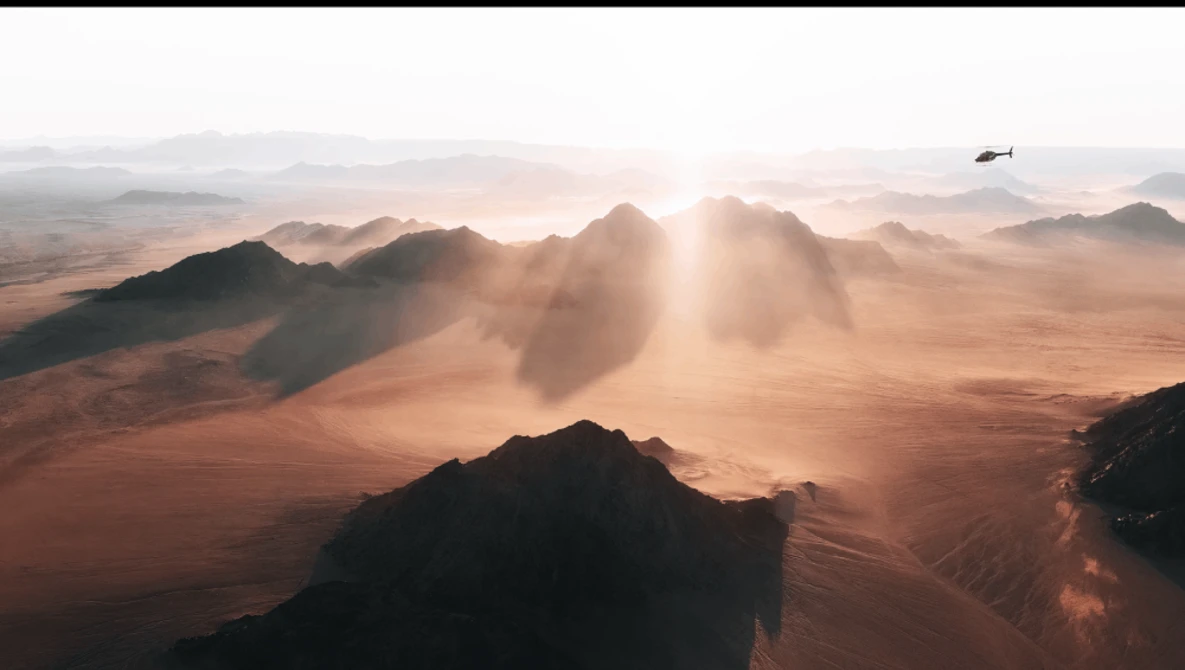 Aerial view of a desert landscape with mountains silhouetted against golden sunlight and dust clouds.