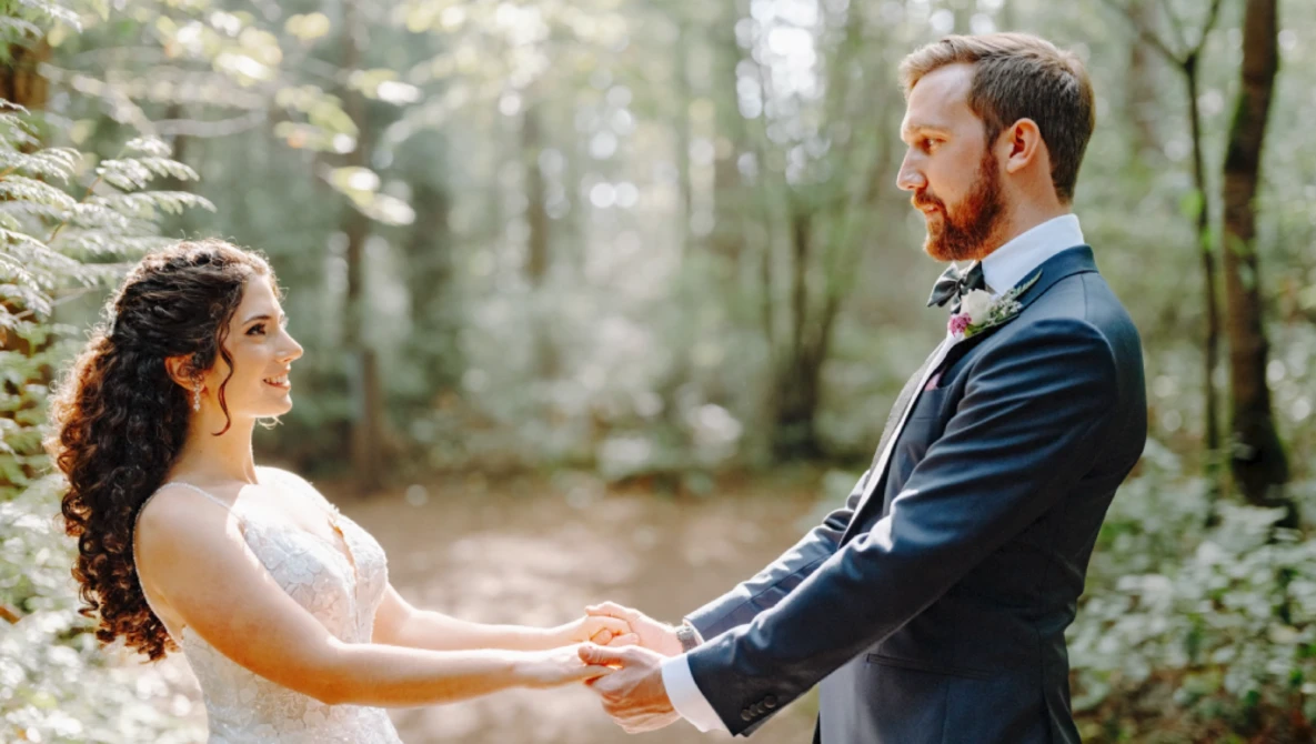 Bride and groom holding hands facing each other in a forest setting.