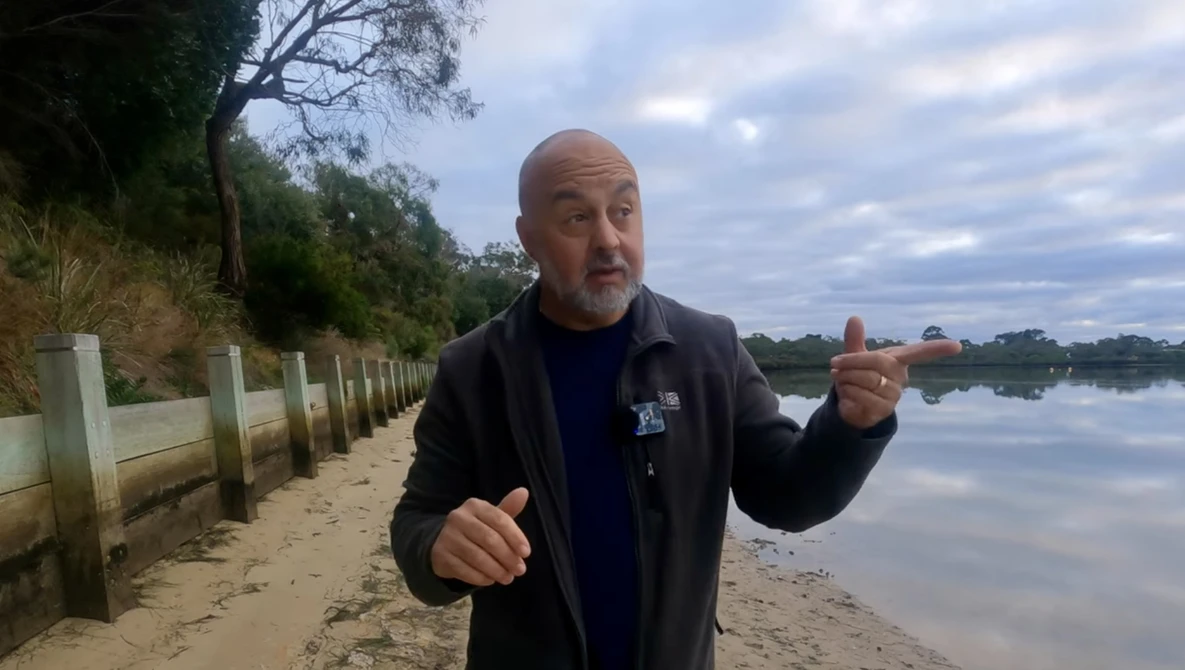 Man gesturing while speaking on a sandy beach beside calm water.