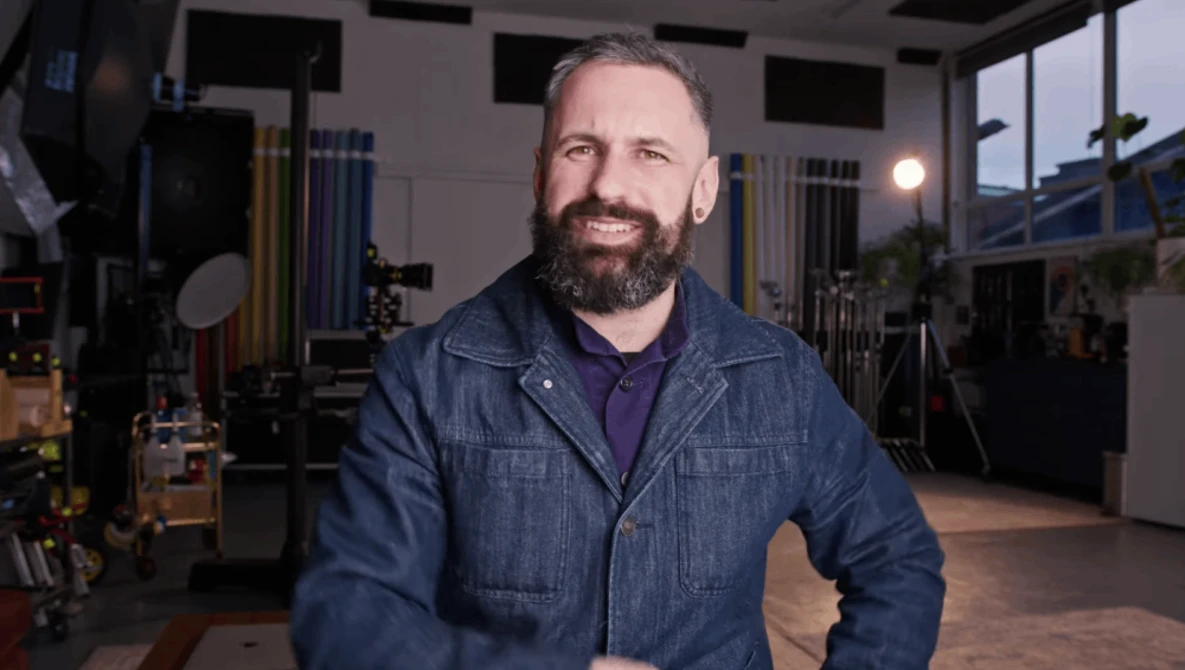 Bearded man in denim jacket smiling in a professional studio setting with lighting equipment visible.