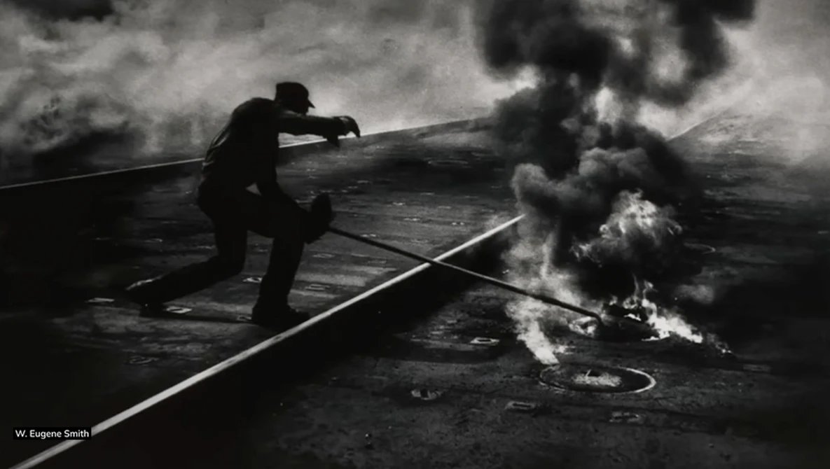 Silhouetted firefighter directing water stream at industrial fire in black and white photograph.