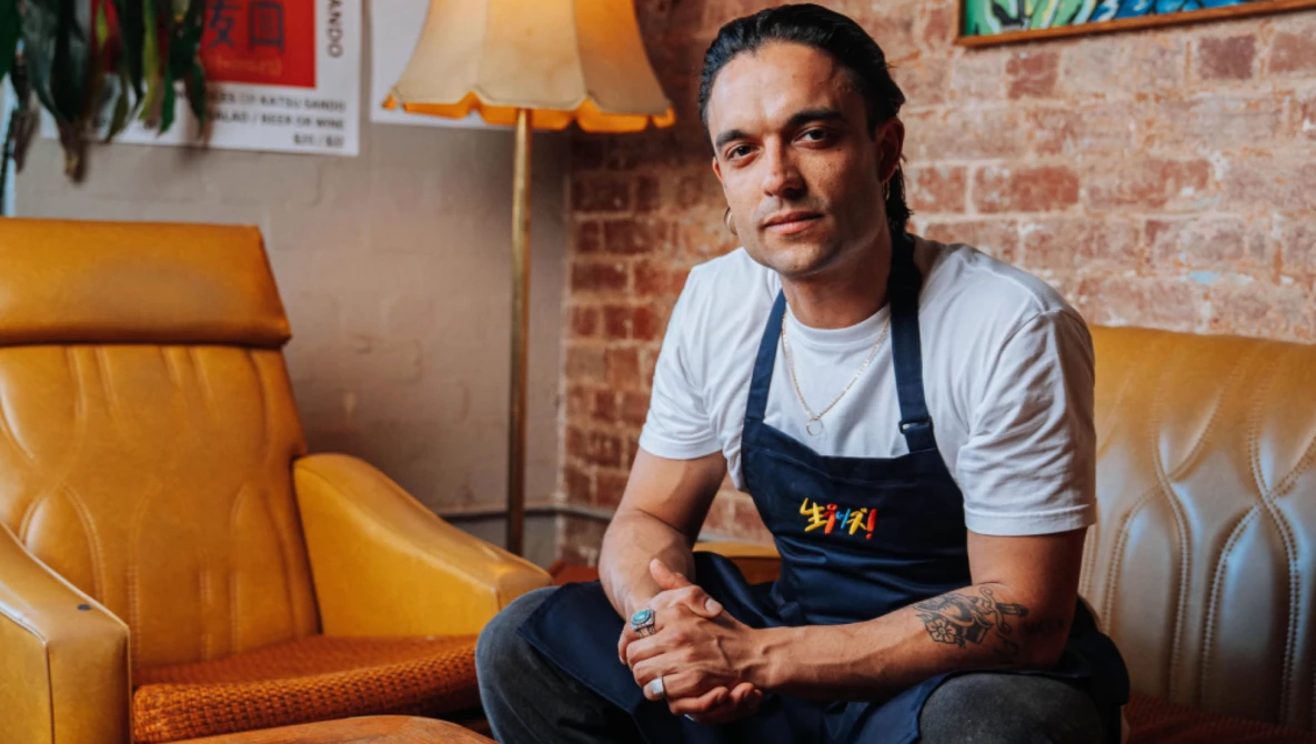 Man in dark apron seated on mustard-yellow leather chair against exposed brick wall.