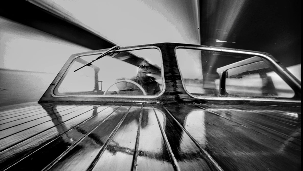 Black and white photo taken from inside a moving car, showing the windshield and interior with motion blur streaking across the frame.
