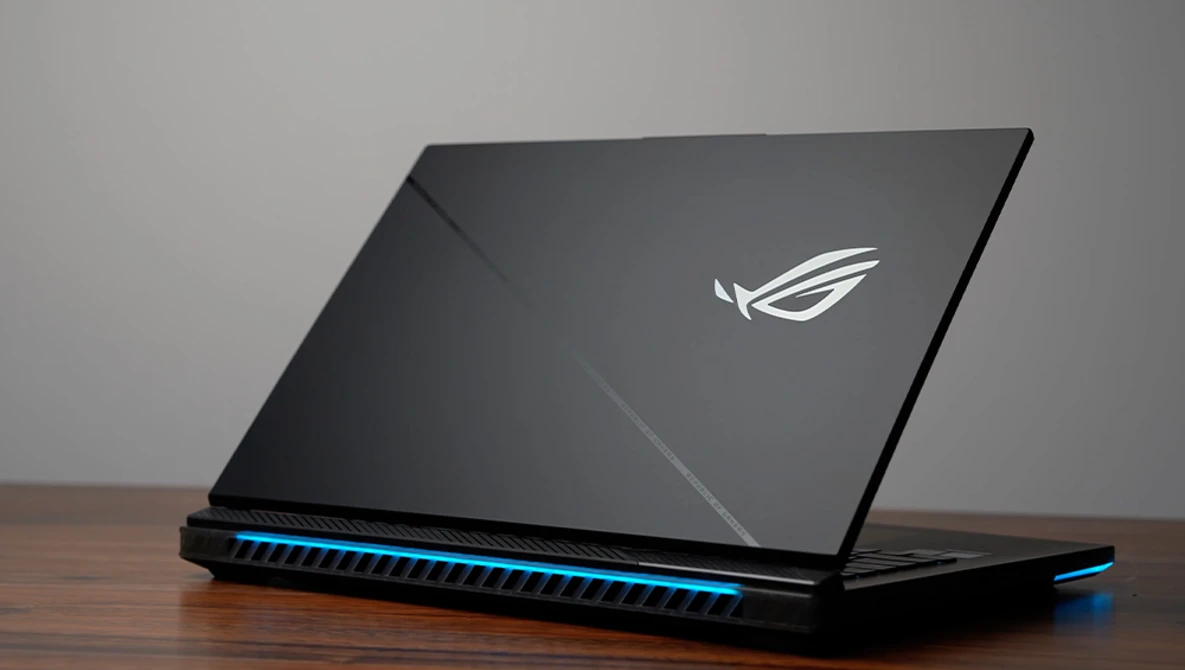 Gaming laptop with illuminated blue accents and winged logo on dark lid, photographed at an angle on wooden surface.