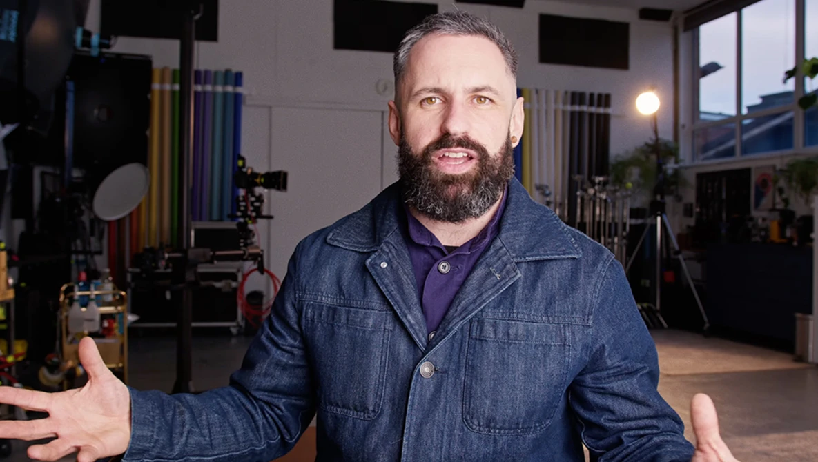 Man with beard in denim jacket gesturing in a professional studio with lighting equipment visible in background.
