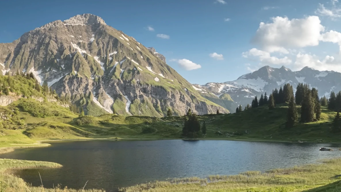 Alpine lake surrounded by coniferous forest with dramatic rocky mountain peak and green meadow.