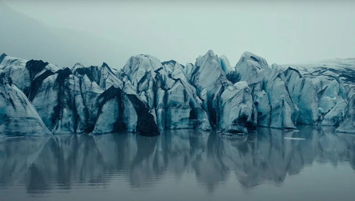 Jagged glacier face with dark crevasses reflected in still water below.