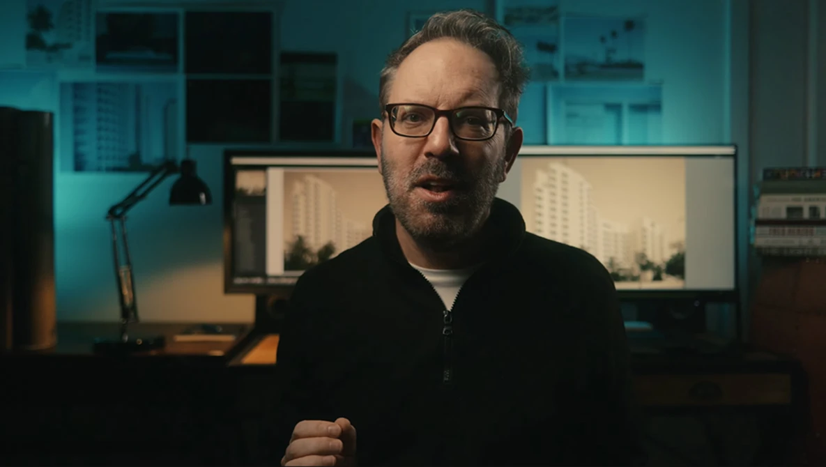 Man with glasses speaking in a studio with dual monitors and teal-blue ambient lighting.