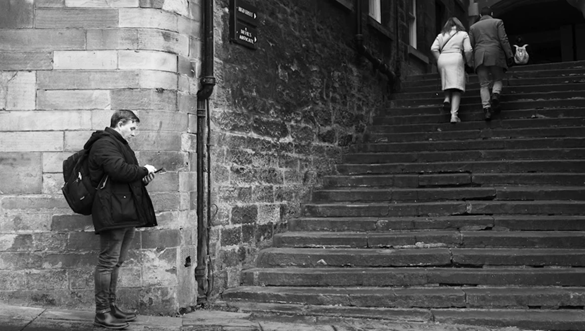 Woman in dark coat standing alone against stone wall while two people ascend steep stone steps in the background.
