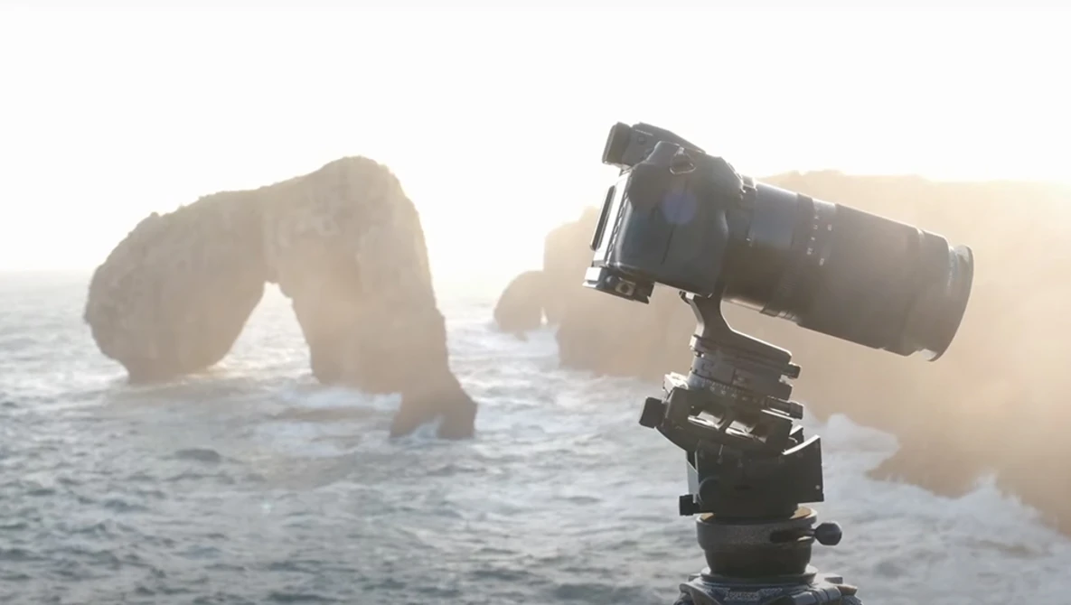 Camera with telephoto lens mounted on tripod head, positioned to photograph large rock formations in misty ocean water.