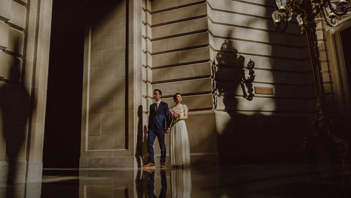 Bride and groom standing in a classical hallway with dramatic side lighting casting shadows on stone walls.