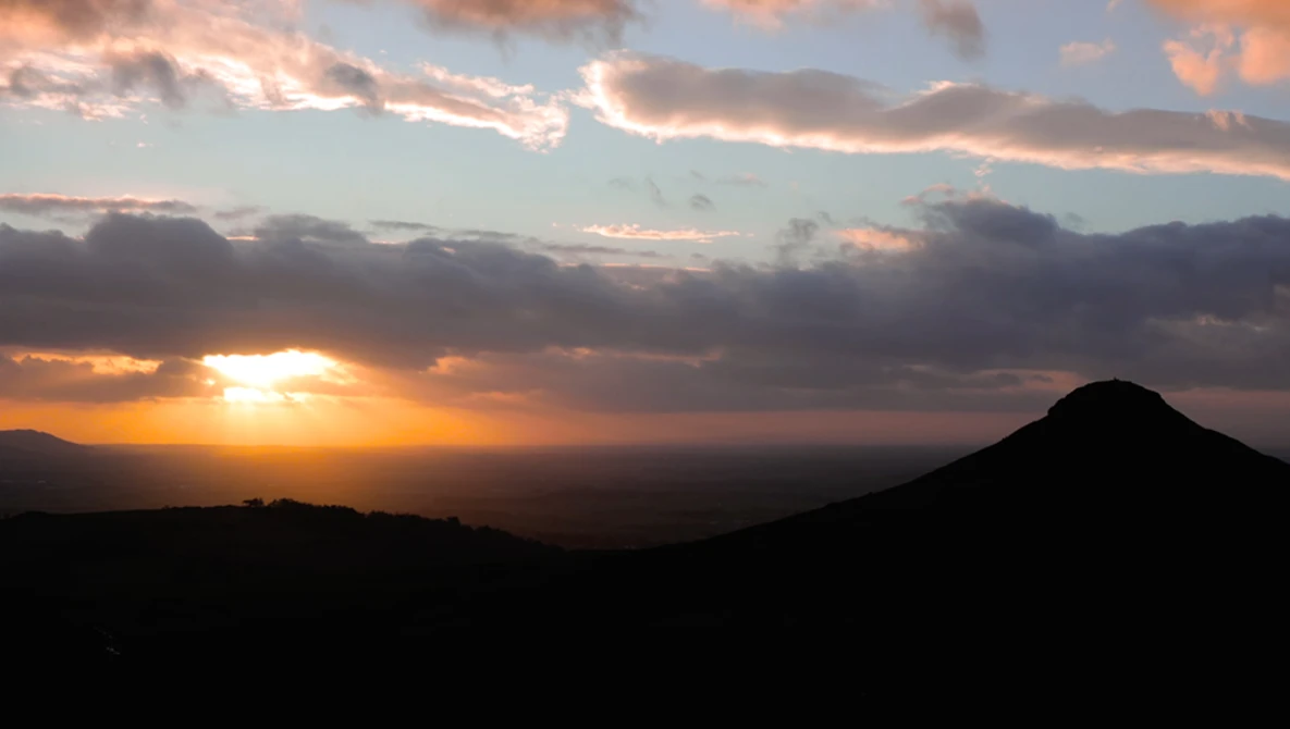 Silhouetted volcanic cone against a golden sunset with dramatic cloud formations and warm orange light breaking through the sky.