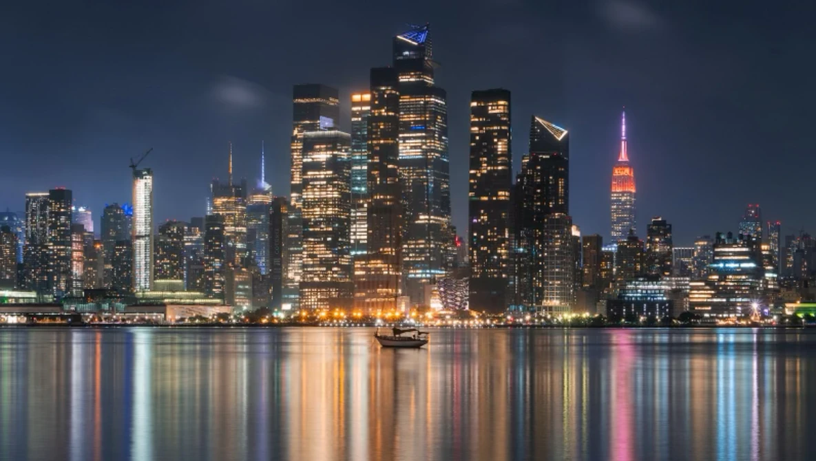 Illuminated Manhattan skyline at dusk reflected in calm river water.