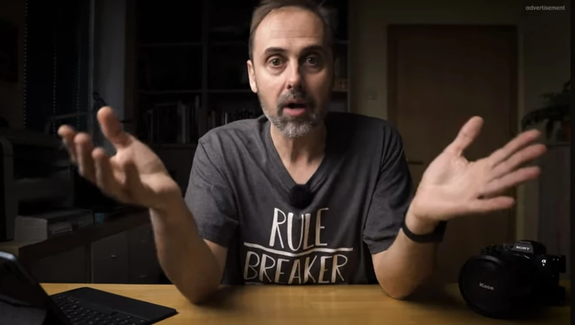 Man in a dark v-neck shirt gesturing expressively while speaking to camera indoors.