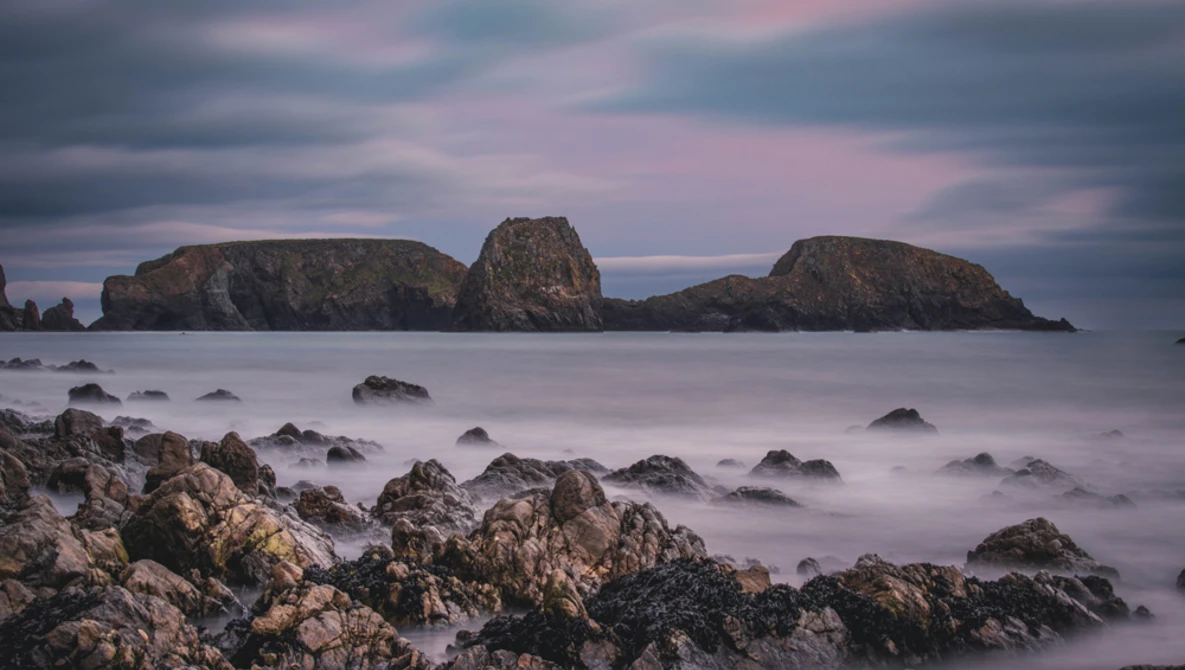 Rocky shoreline with smooth water and three distant islands under a twilight sky with pink and blue clouds.