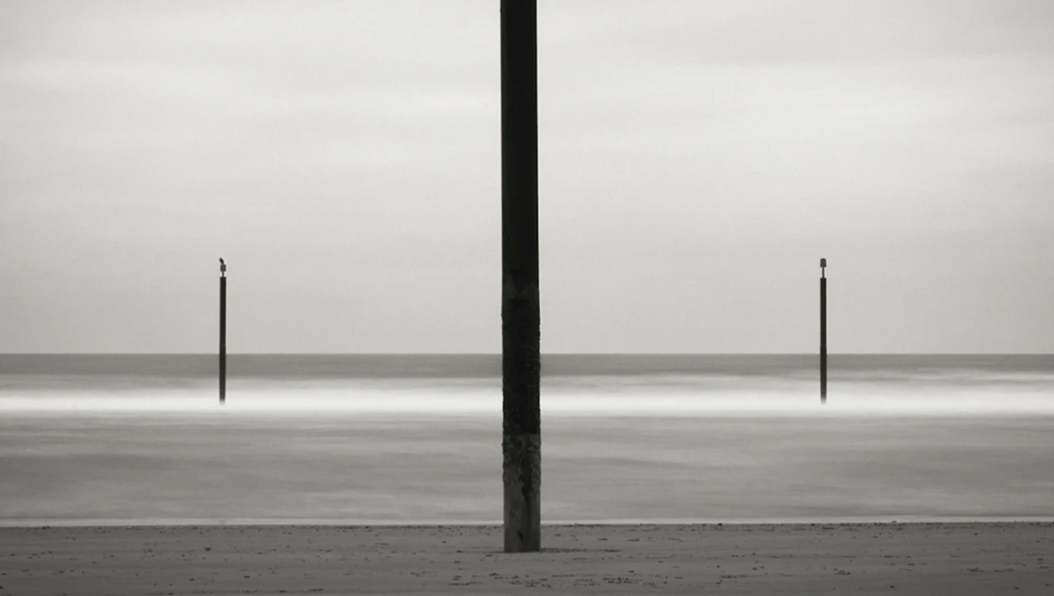 Three vertical posts on a vast, flat beach with calm water and overcast sky in stark monochromatic composition.