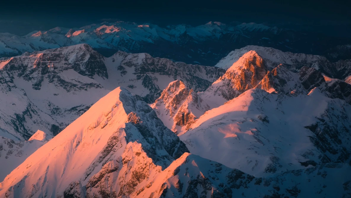 Snow-covered mountain peaks illuminated by golden alpenglow at sunset or sunrise.